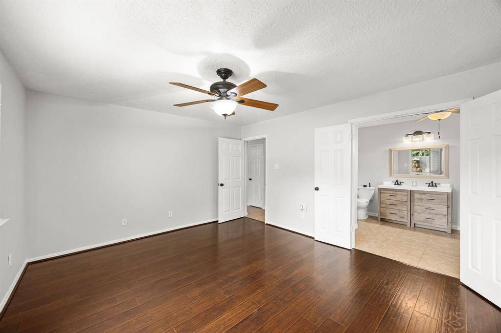 This spacious primary bedroom features elegant dark wood look flooring, a ceiling fan, and neutral walls. It opens to a modern en-suite bathroom with double sinks and a large mirror, creating a comfortable and stylish retreat.