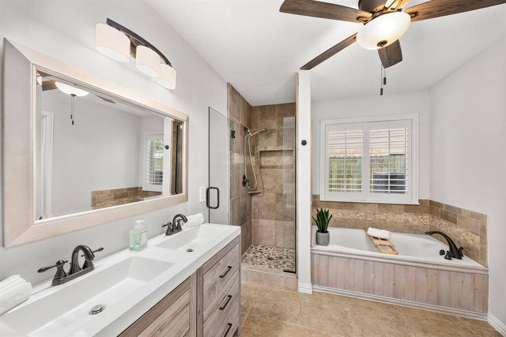 This primary bathroom features a modern double vanity with black fixtures, a spacious walk-in shower with glass doors, a soaking tub with tile accents, and a ceiling fan. Natural light streams in through a window with shutters, highlighting the neutral tones and clean design.