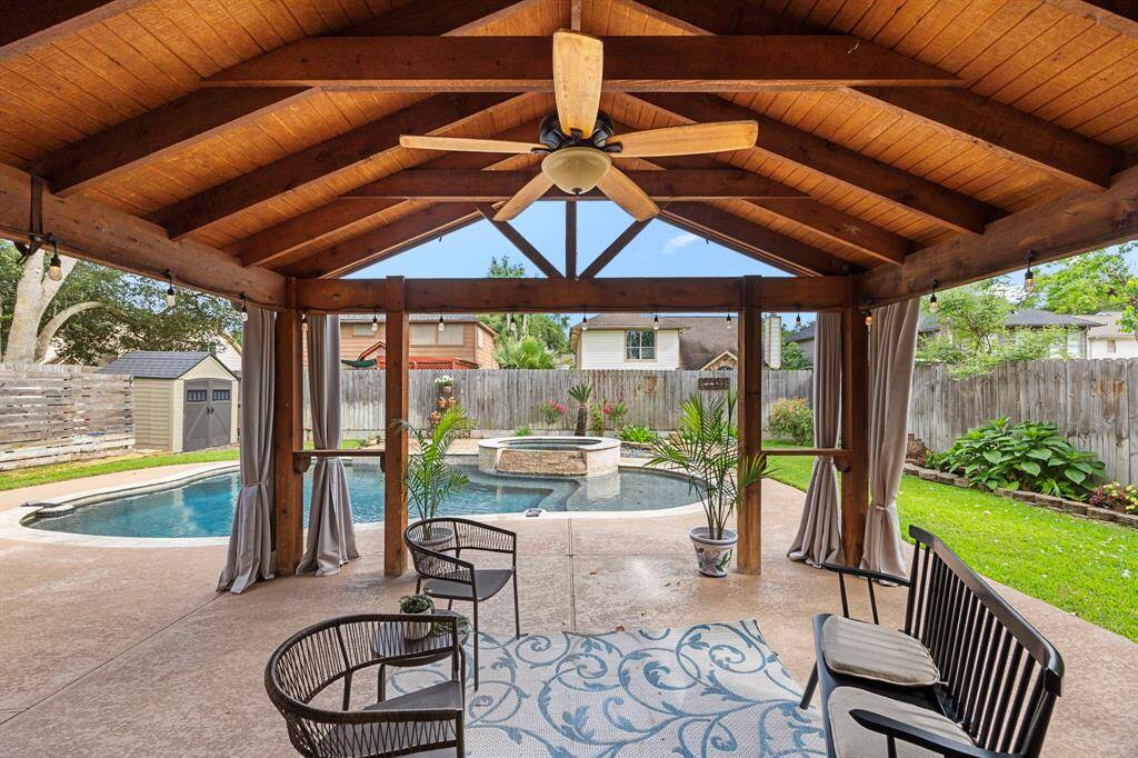This backyard features a spacious covered patio with a wooden ceiling and a ceiling fan, overlooking a well-maintained pool. The area includes comfortable seating, lush greenery, and privacy fencing, offering a perfect space for relaxation and entertainment.