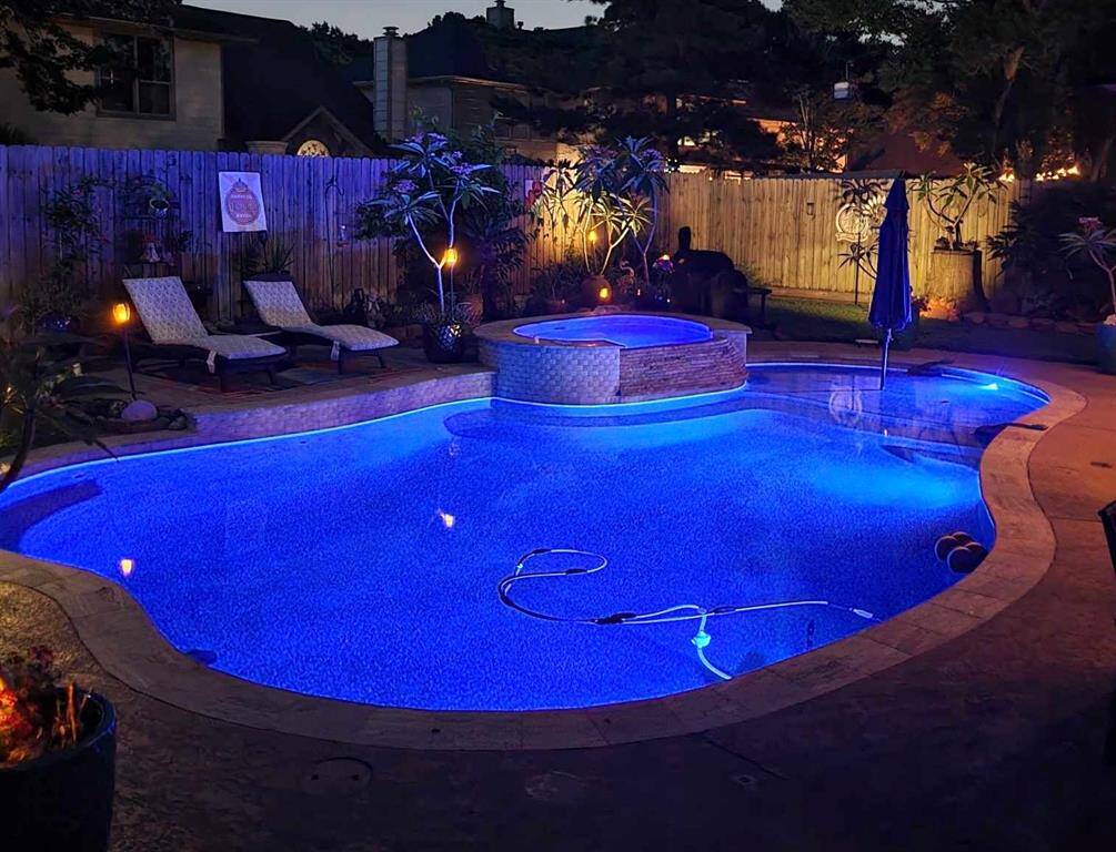 This photo showcases a beautifully lit backyard pool area at night, featuring a freeform swimming pool with a connected hot tub. The space is surrounded by a wooden privacy fence, lush plants, and includes lounge areas for relaxation, creating a serene and inviting outdoor oasis.