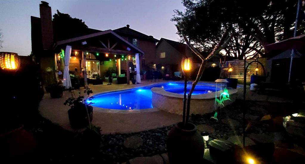 This photo showcases a cozy backyard at dusk featuring a well-lit pool with an adjoining spa. A covered patio with ambient lighting and outdoor seating area creates an inviting space for relaxation and entertainment. Surrounded by mature trees and decorative lanterns, it offers a serene and private atmosphere.