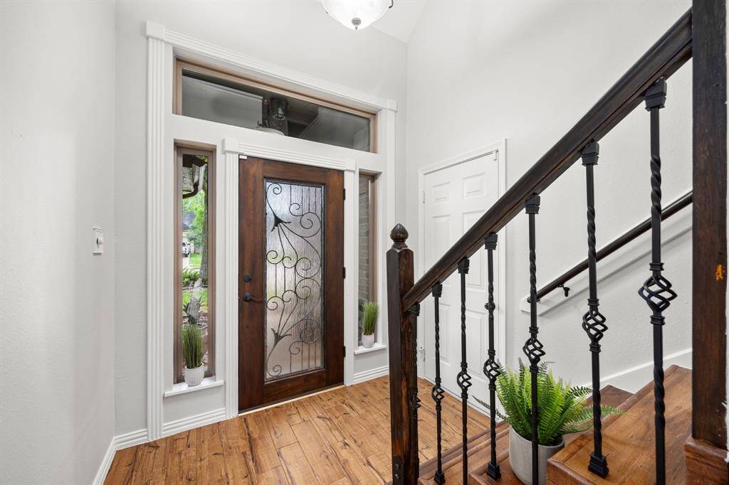 The photo showcases a bright, welcoming entryway featuring a wooden front door with decorative glass panels, flanked by tall windows. The space has warm wood flooring and a staircase with elegant wrought iron railings.