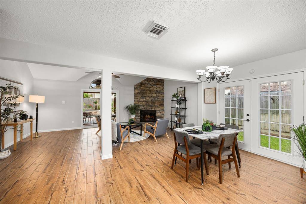 This inviting open-concept living space features warm wood look tile flooring, a cozy stone fireplace, and abundant natural light from large windows and French doors leading to a patio. A dining area with a modern chandelier enhances the home's charm and functionality.