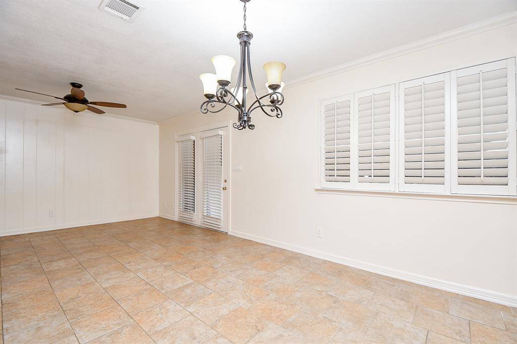 This room features tiled flooring, a ceiling fan, and a stylish light fixture. It has white walls with large windows and French doors, both with plantation shutters, offering a bright and airy feel.