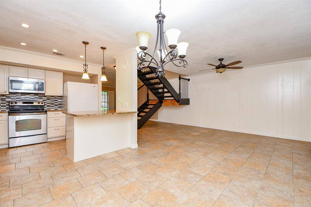 This photo showcases an open-concept living and kitchen area with tile flooring. The kitchen features stainless steel appliances, a stylish backsplash, and pendant lighting above a breakfast bar. A modern staircase leads to an upper level, and the room is well-lit with a ceiling fan and elegant chandelier.