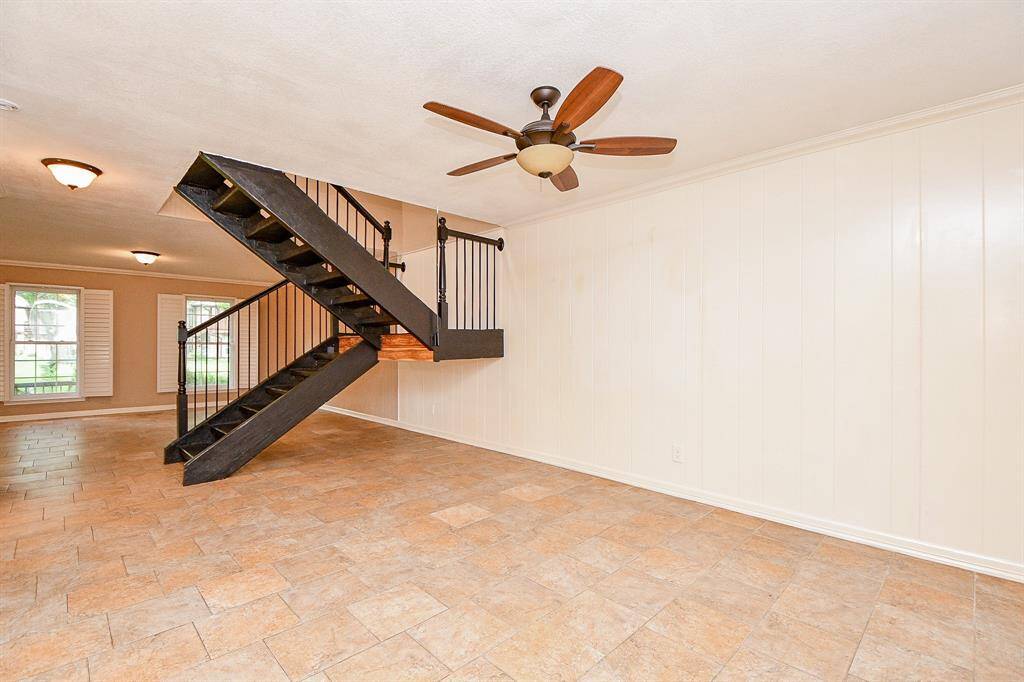 Spacious living area with a modern floating staircase, tile flooring, and a ceiling fan. Large windows with shutters provide natural light, creating a bright and airy atmosphere.