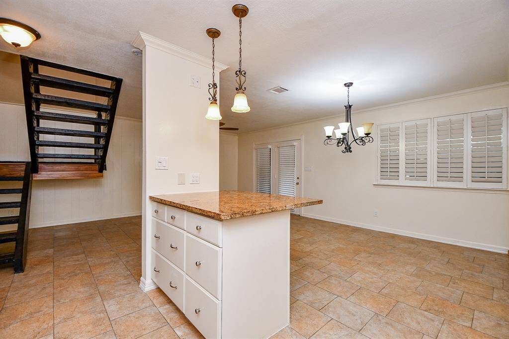 This photo showcases a spacious, open-concept living area with tiled flooring and a modern staircase. It features a kitchen island with granite countertops and stylish pendant lighting, alongside large windows with plantation shutters for ample natural light.
