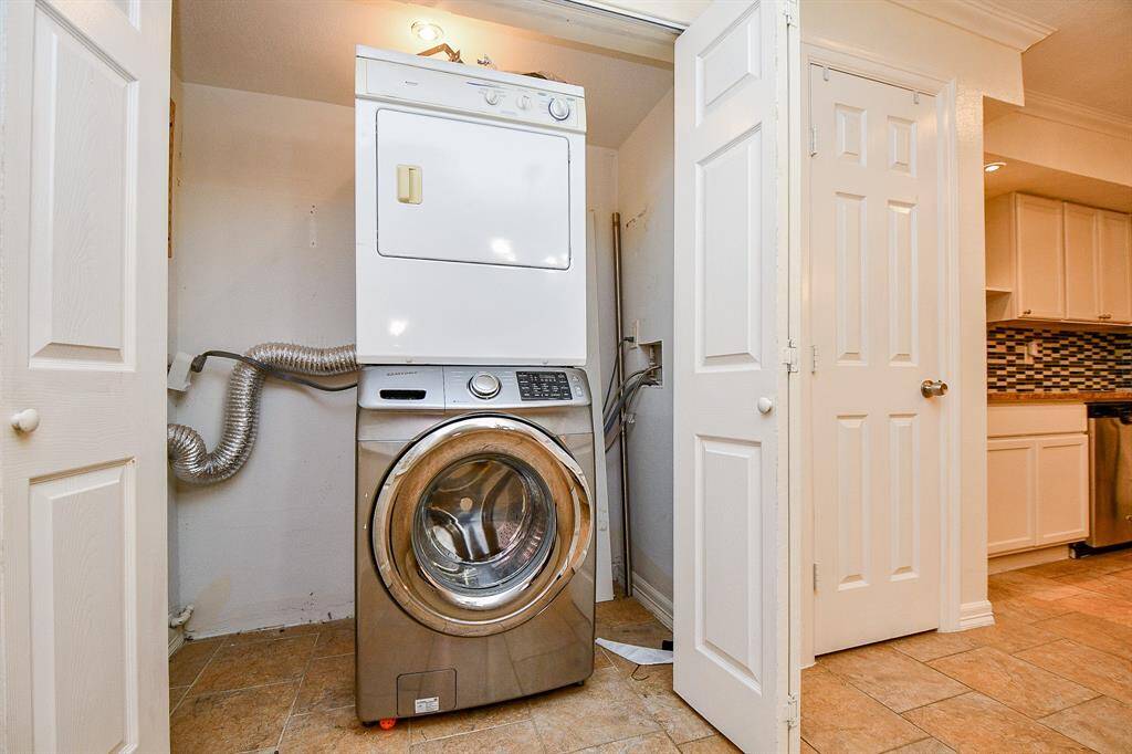 This photo shows a compact laundry area with a stacked washer and dryer in a closet, featuring tiled flooring and adjacent to a kitchen with modern cabinetry and a tiled backsplash.