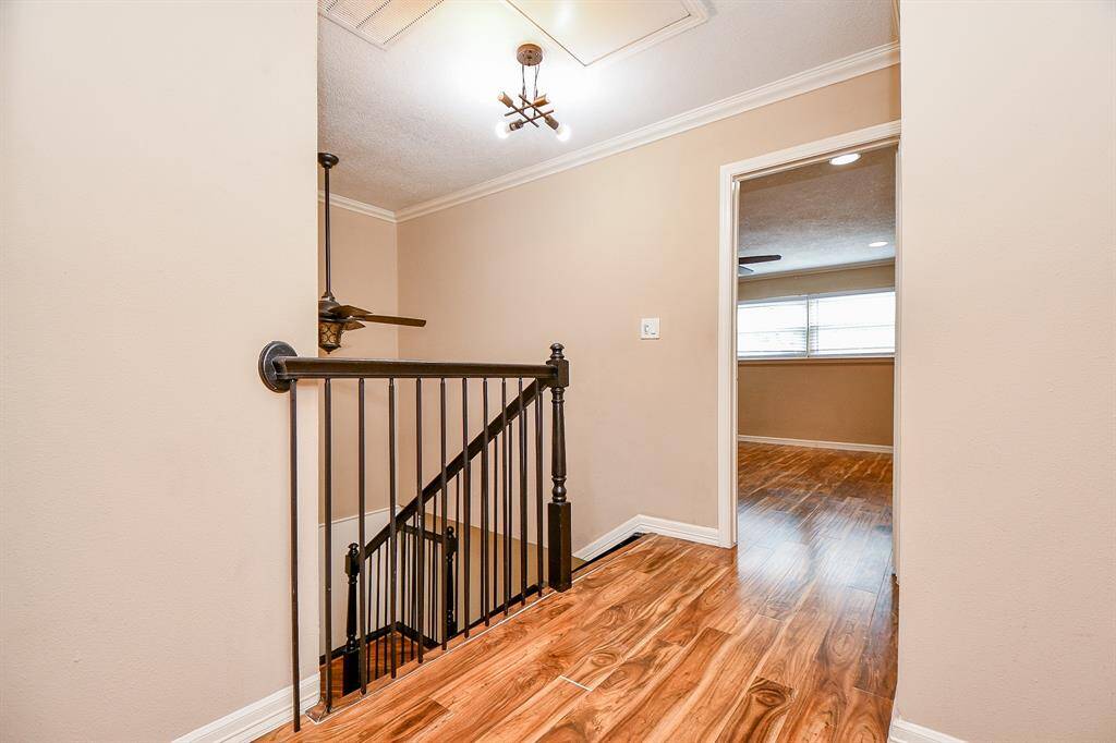 This photo shows a bright hallway with wooden flooring and a metal railing leading to a staircase. The walls are painted in a neutral tone, and there's a ceiling light fixture. An adjacent room with large windows and similar flooring is visible.