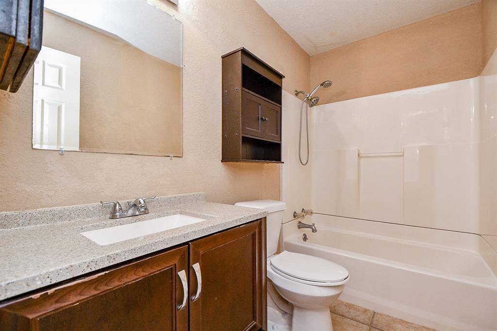 This bathroom features a modern vanity with a granite countertop, a large mirror, and a combination shower-tub. It includes a mounted storage cabinet above the toilet and has neutral-toned walls and tile flooring.