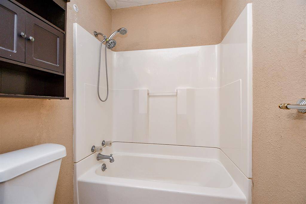 This bathroom features a clean, white bathtub with a showerhead, enclosed by a simple surround. There's a dark wood storage cabinet above the toilet, and the walls are painted in a neutral beige tone.