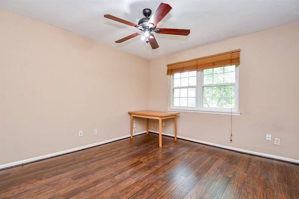 This room features warm wood flooring, neutral walls, and a ceiling fan. A large window with wooden blinds offers ample natural light, and there's a small wooden table in the corner.
