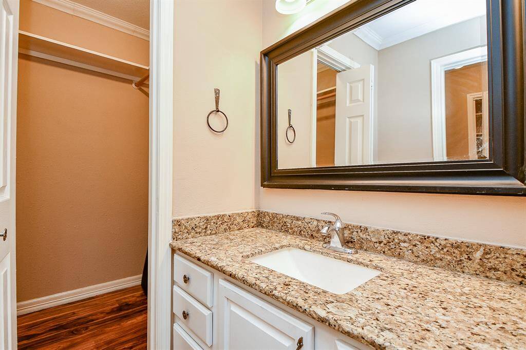 This photo showcases a bathroom with a granite countertop and an undermount sink. It features a large framed mirror, white cabinetry, and an adjacent walk-in closet with wood flooring.