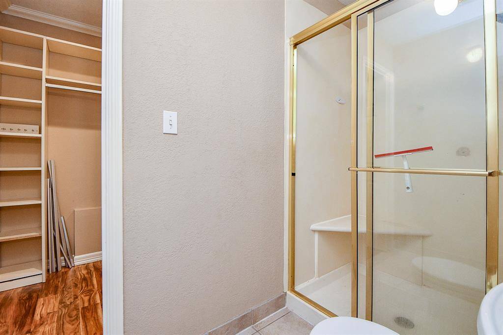 This photo shows a bathroom with a glass-enclosed shower featuring gold trim. Adjacent is a walk-in closet with wooden shelving and laminate flooring. The space is compact and functional.