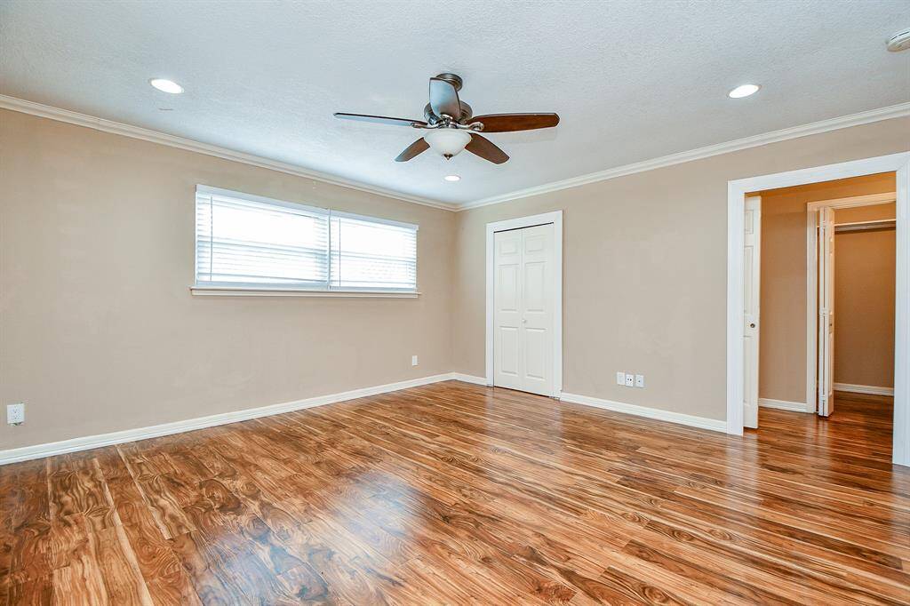 This spacious room features polished hardwood floors, neutral walls, and recessed lighting. It includes a ceiling fan, a large window for natural light, and access to a closet and an adjoining space.