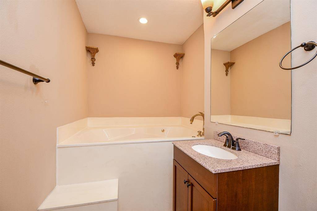 This bathroom features a cozy corner tub with elegant fixtures, a single-sink vanity with a granite countertop, and warm-toned walls. Ideal for relaxation.