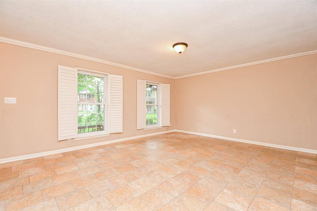 This spacious, light-filled room features neutral walls, tiled flooring, and two large windows with shutters, offering a bright and inviting atmosphere.