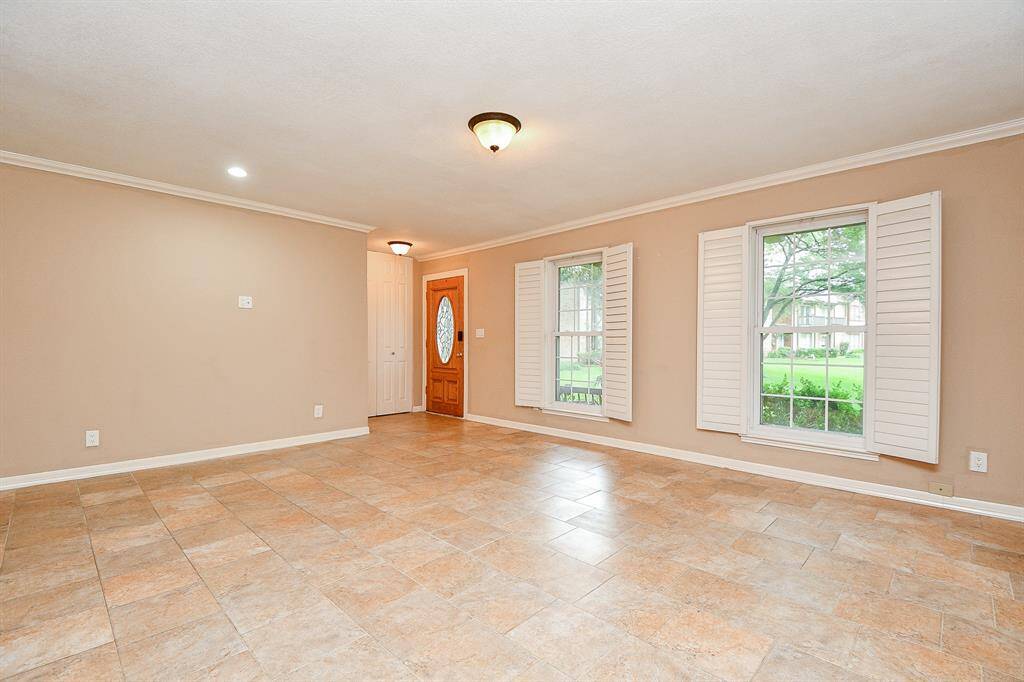 Bright living area with tiled floors, neutral walls, and crown molding. Features large windows with shutters and a wooden front door. Perfect for natural light and open space.