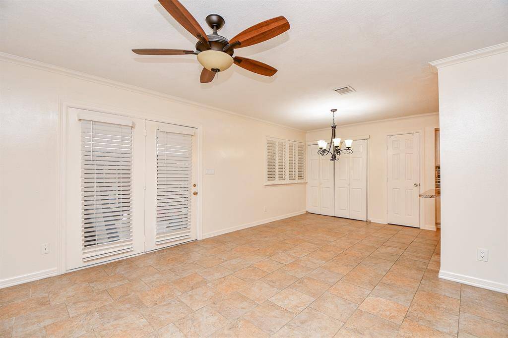 This spacious room features tile flooring, a ceiling fan, and a chandelier. It includes French doors with blinds leading outside, and an additional window with plantation shutters for ample natural light. Two interior doors offer access to adjacent spaces.