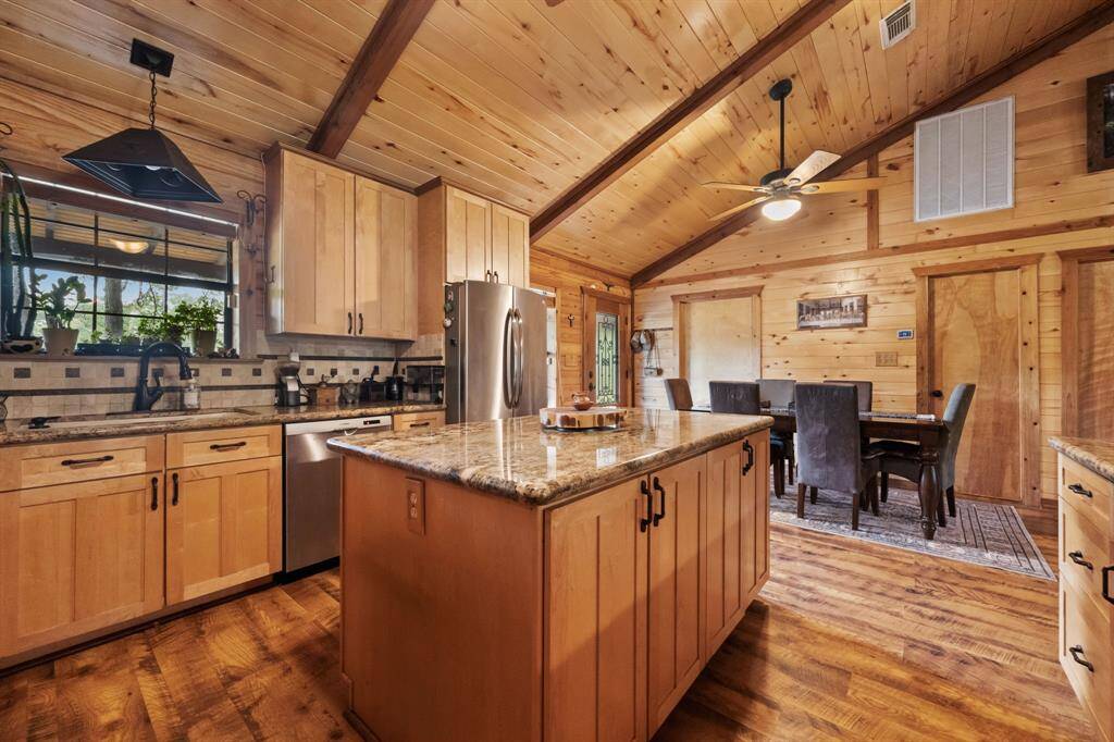 A warm, inviting kitchen and dining area with a rustic feel. It features wooden cabinetry, granite countertops, and a spacious island. The vaulted ceiling with exposed beams and ceiling fan adds character, while the dining area provides a cozy space for meals. Natural light enhances the wooden interior, creating a charming atmosphere.
