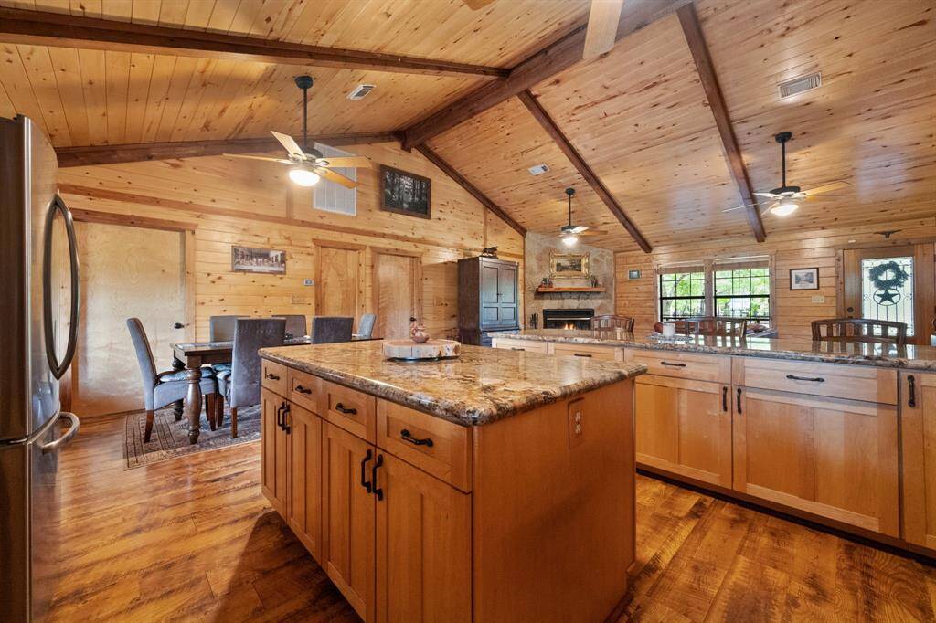 This inviting kitchen and dining area features warm wooden interiors with vaulted ceilings and exposed beams. It includes a large island with granite countertops, ample cabinetry, and modern appliances. The space is open and airy, perfect for entertaining, with a cozy fireplace adding charm.