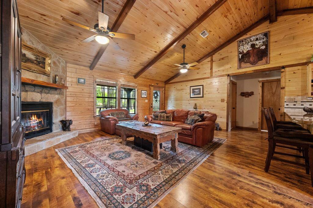 This cozy living room features a high vaulted ceiling with exposed wooden beams, a stone fireplace, and hardwood flooring. It includes a warm, rustic design with wood-panels.