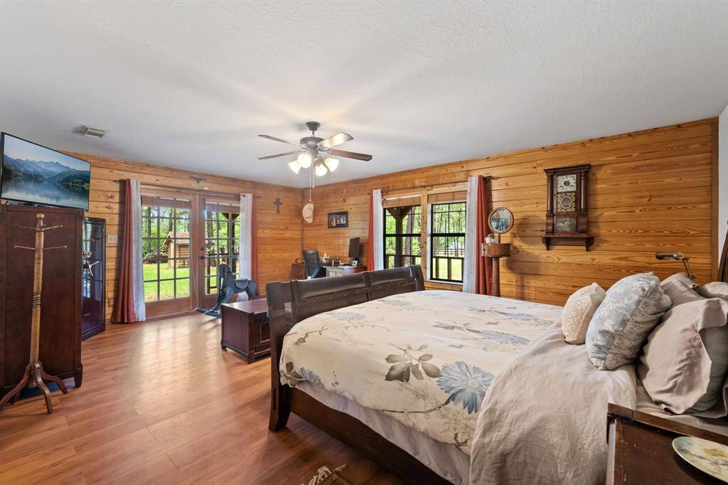 This cozy bedroom features wood-paneled walls and flooring, creating a rustic feel. It has ample natural light from large windows and French doors leading to the outside. The room includes a ceiling fan, wall-mounted TV, and a comfortable bed, perfect for relaxation.