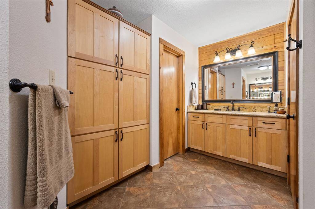 This bathroom features warm wooden cabinetry and accents, a spacious vanity with dual sinks, and a large mirror. The tile flooring and ample storage enhance functionality, creating an inviting and practical space.