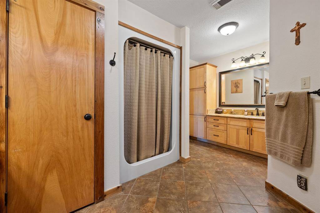 This bathroom features a stand up shower with a curtain, warm wooden cabinetry, and a spacious vanity with a large mirror. The tiled floor and soft lighting create a welcoming atmosphere.