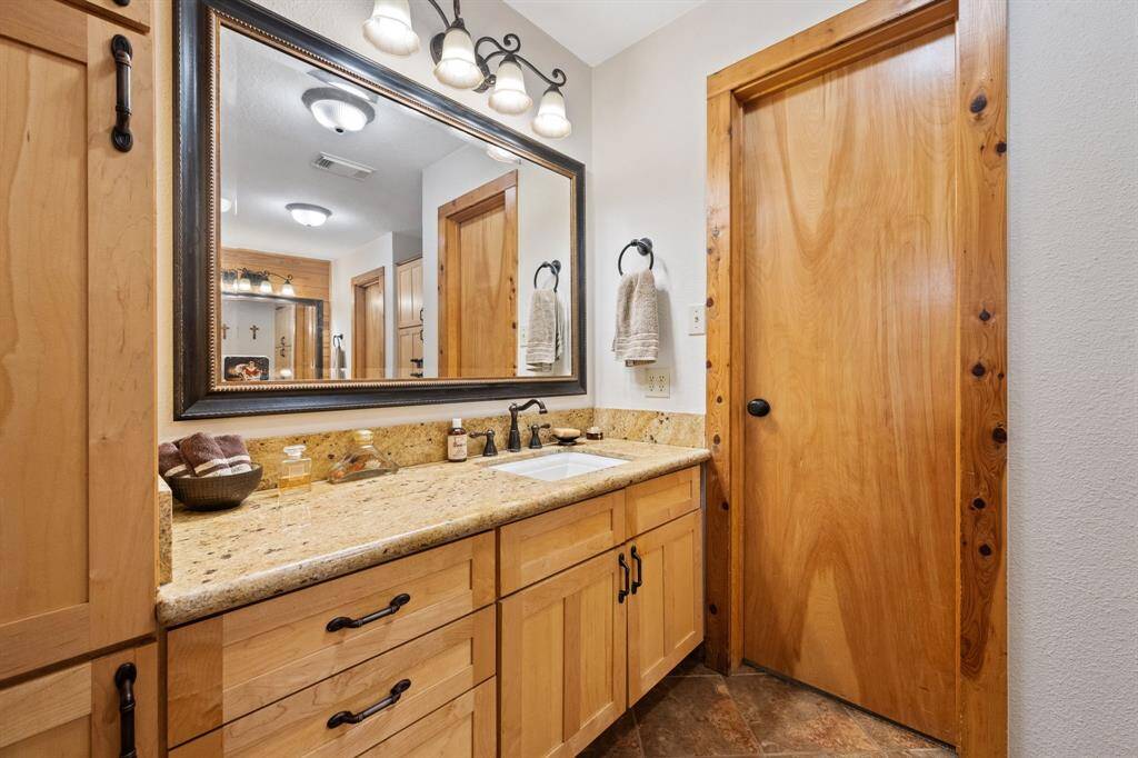 This bathroom features a warm, rustic design with wooden cabinetry and door, a granite countertop, and a large framed mirror. Modern light fixtures and neutral tones create a cozy, inviting atmosphere.