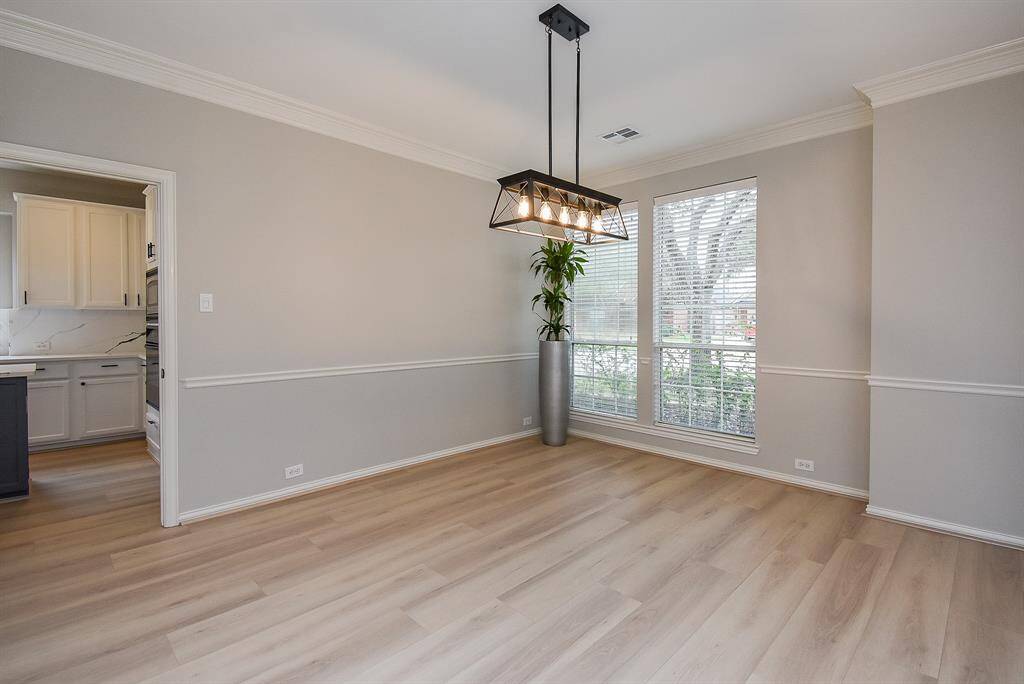 This photo showcases a bright dining room with light flooring, neutral walls, and a modern rectangular chandelier. Large windows offer ample natural light. An opening leads to a kitchen with white cabinetry.