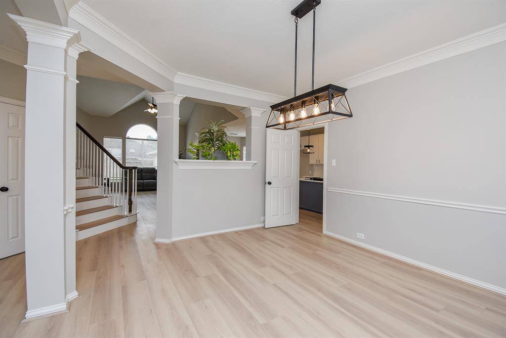 This photo showcases a bright, open living space with a modern chandelier, light wood flooring, and elegant crown molding. There is a staircase leading to an upper level and a partial view into a cozy living area with large windows. A doorway leads to a kitchen or dining area.