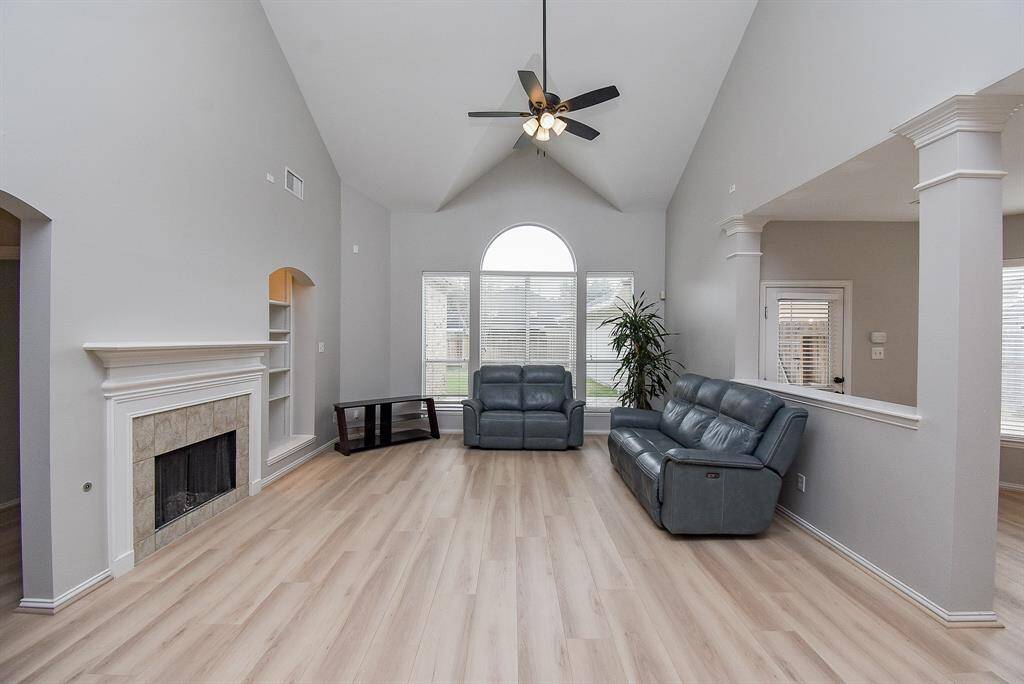 This spacious living room features high vaulted ceilings and abundant natural light from large windows. It includes a cozy fireplace with a modern mantel, built-in shelving, and elegant light flooring. The area is perfect for relaxation with ample seating.