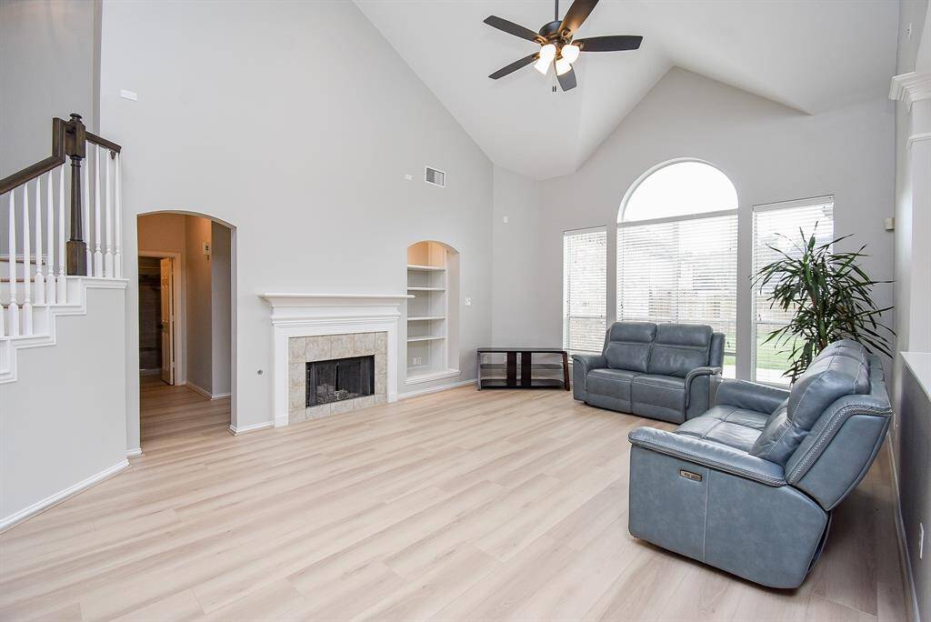 This spacious living room features high vaulted ceilings, large arched windows for natural light, a cozy fireplace, and modern light flooring. Comfortable seating and a neutral color palette create a welcoming atmosphere.