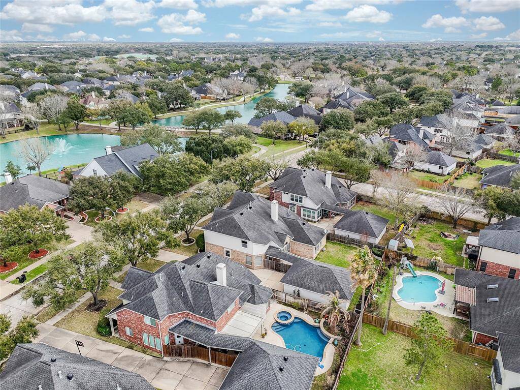 This aerial photo showcases a suburban neighborhood with spacious homes, tree-lined streets, and a scenic view of a nearby lake with fountains. The property features a private backyard, offering a relaxing outdoor space. Ideal for families seeking a tranquil, community-oriented setting.