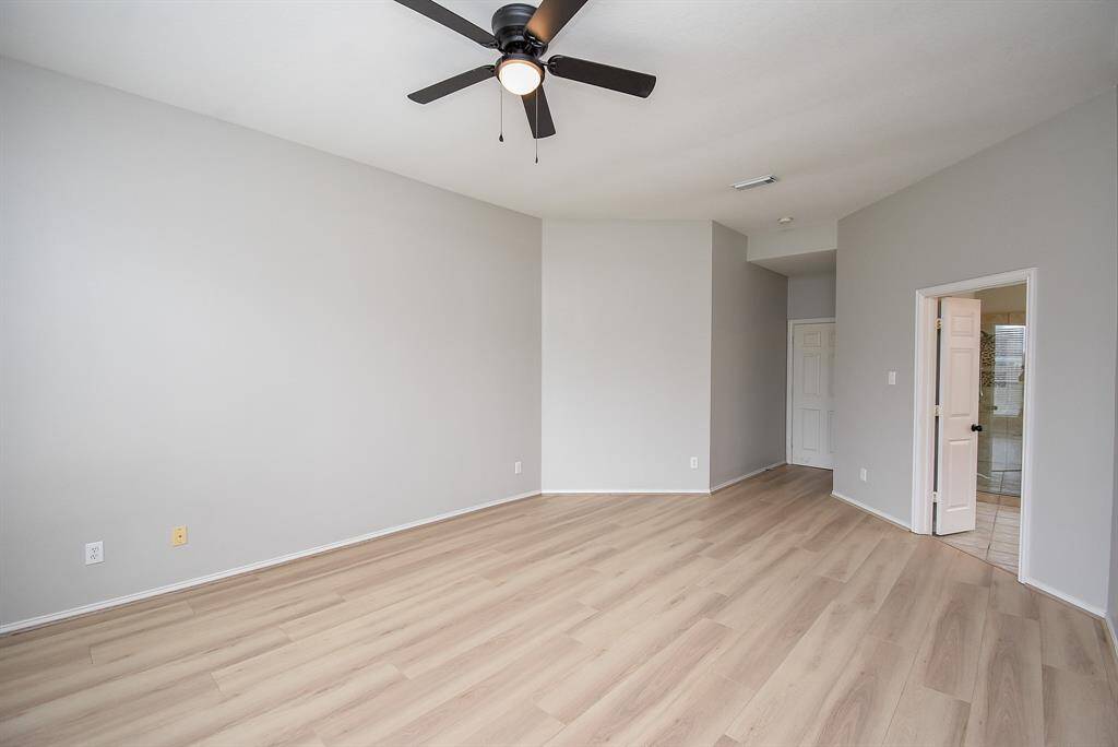 This primary bedroom features a modern ceiling fan, light flooring, and neutral walls. It opens to a hallway and a bathroom, offering a spacious and versatile layout.