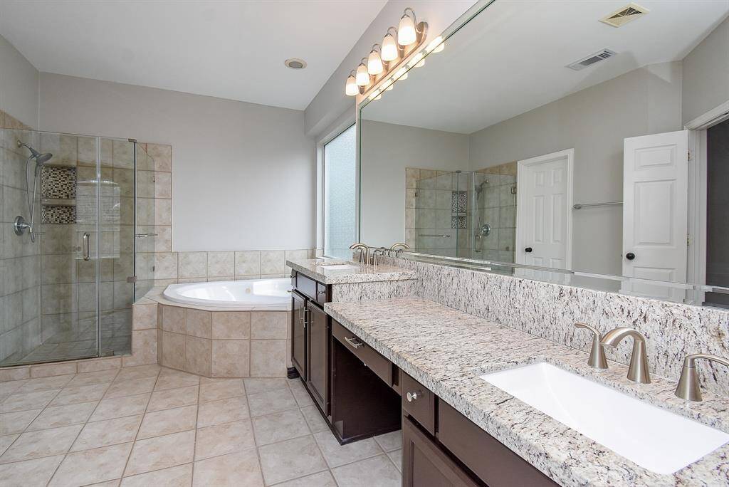 This spacious primary bathroom features a double sink vanity with granite countertops, a luxurious glass-enclosed shower, and a relaxing soaking tub. The neutral tile flooring and ample natural light create a serene and inviting atmosphere.