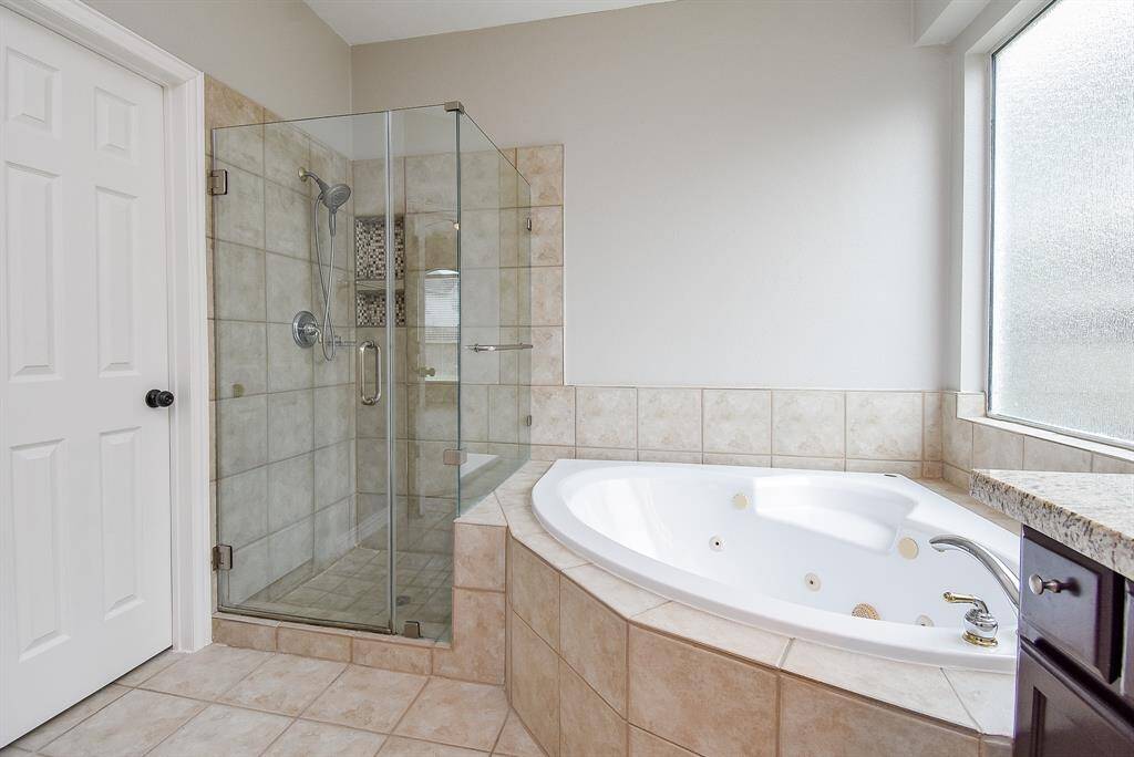 This bathroom features a spacious corner jetted bathtub and a glass-enclosed shower. The neutral tile design and large frosted window provide a bright and elegant atmosphere.