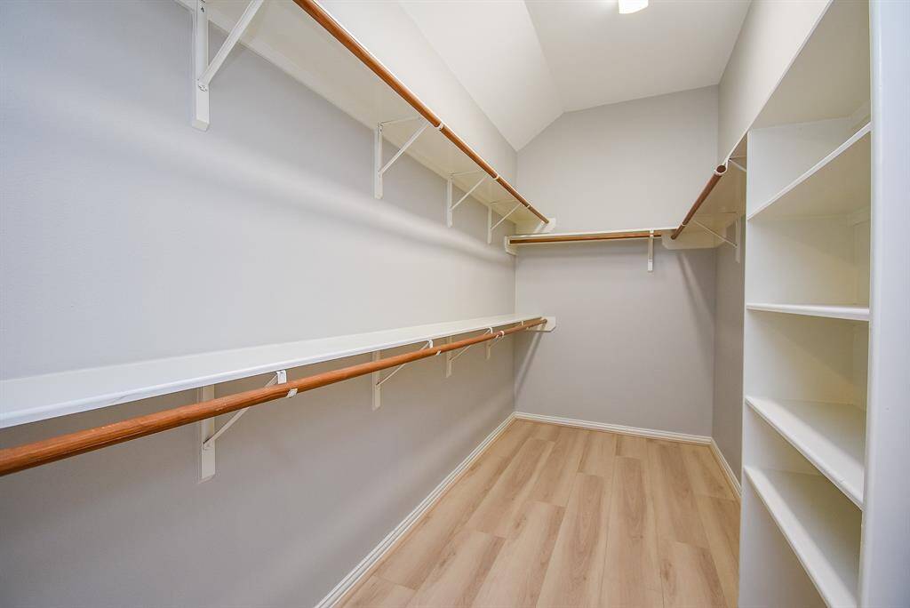 This photo shows a spacious walk-in closet with light flooring, ample hanging space, and built-in shelving, offering plenty of storage options.