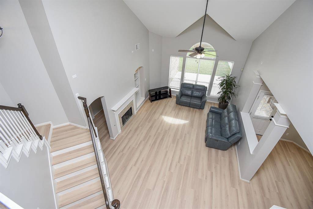 This spacious living room features high vaulted ceilings, light flooring, and abundant natural light from large windows. It includes a cozy fireplace, modern ceiling fan, and an open-concept layout with a staircase leading up.