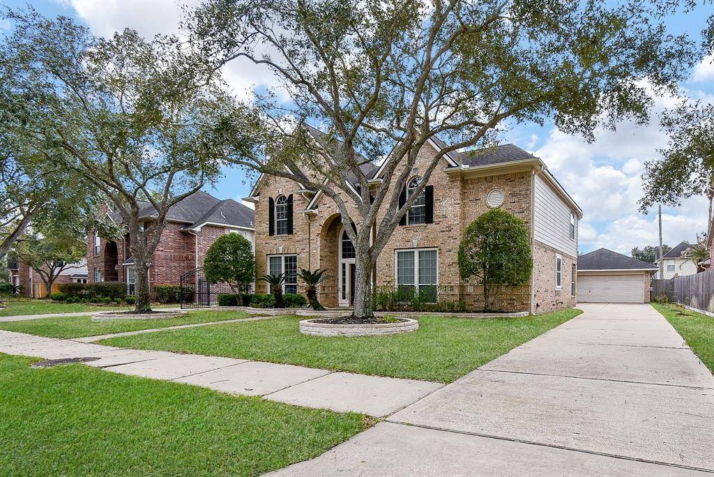 This photo showcases a two-story brick home with a classic design. It features a spacious driveway leading to a detached garage, and a well-maintained front yard with mature trees. The house has a welcoming entrance with arched windows and black shutters.