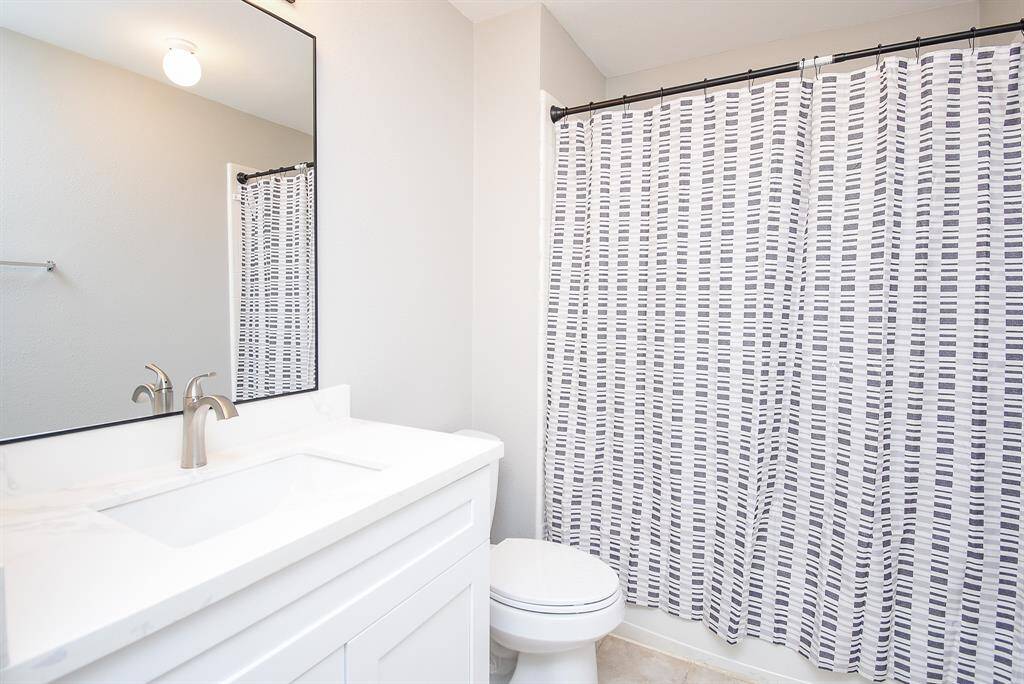 This bathroom features a modern design with a white vanity, sleek faucet, large mirror, and tiled flooring. It includes a shower with a stylish patterned curtain, creating a clean and inviting space.