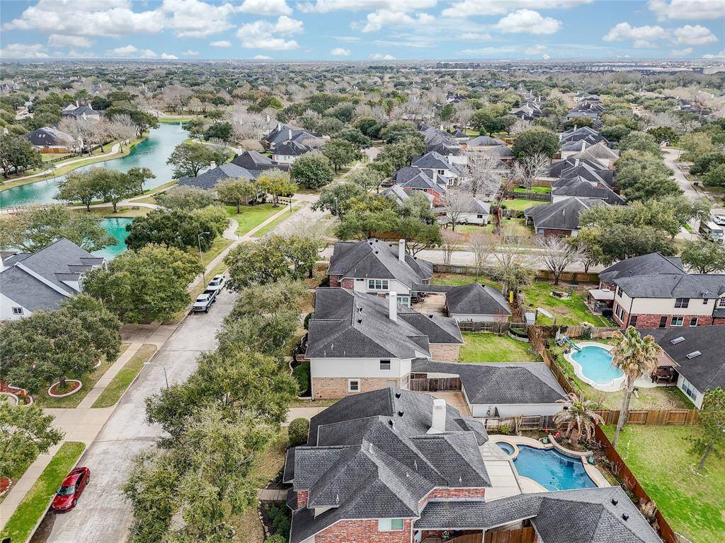 This aerial photo showcases a suburban neighborhood with well-maintained homes, abundant greenery, and a nearby water feature. The area looks peaceful, with tree-lined streets and several homes featuring private pools. Perfect for families seeking a serene community.