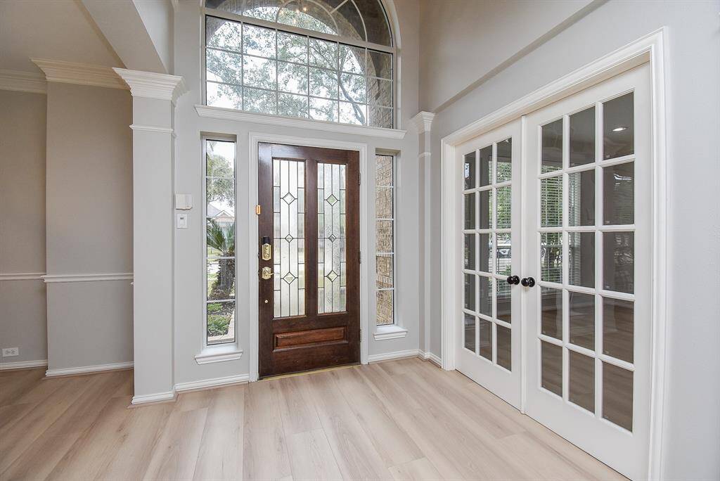 This entryway features a grand wooden door with decorative glass panels, flanked by tall windows. The space is bright and welcoming, with light wood flooring and double glass French doors leading to the study. High ceilings and neutral walls create an open, airy feel.