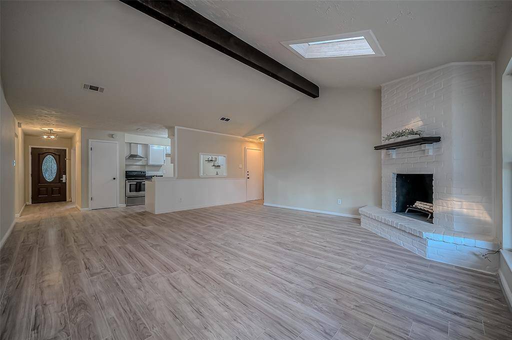 This spacious living area features modern wood-look flooring, a white brick fireplace, high ceilings with exposed beams, and ample natural light. The open-plan layout flows into the kitchen, perfect for entertaining.