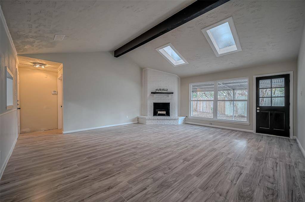 Spacious living room with contemporary gray wood flooring, a white brick fireplace, large new windows offering natural light, and a glass-paned door leading outside. The room features a high ceiling with exposed dark beams and two skylights.