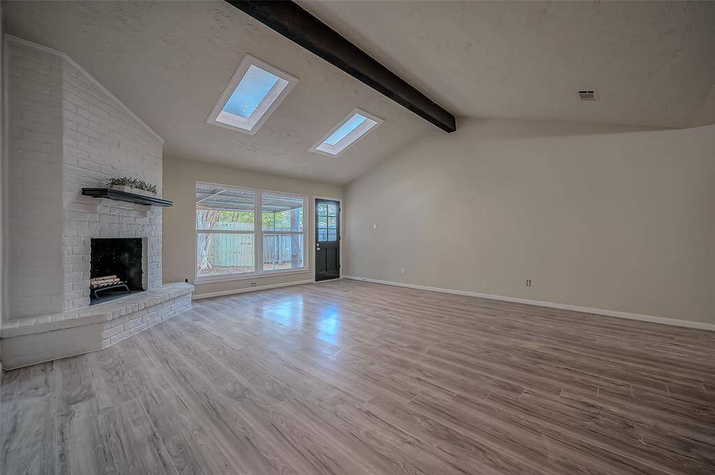 Spacious living area with high ceilings, abundant natural light from skylights and large windows, a white brick fireplace, and modern wood-like flooring.