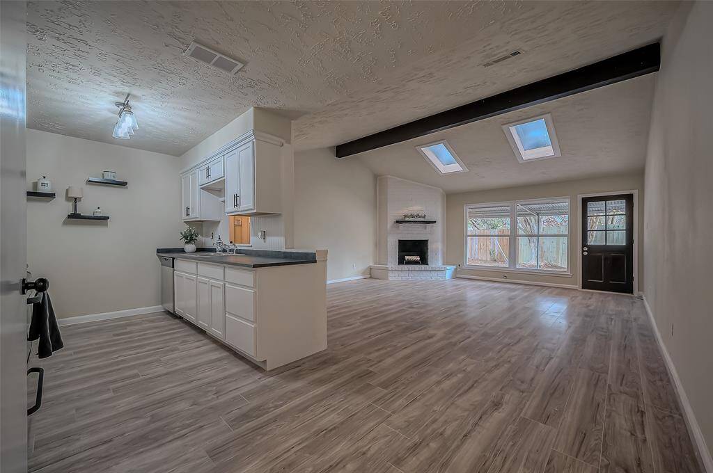 This is a spacious, open-concept living area with a modern kitchen, featuring white cabinetry and a central island. There's ample natural light from skylights and a large window, plus a fireplace adds a cozy touch. The room has contemporary wood-look flooring throughout.