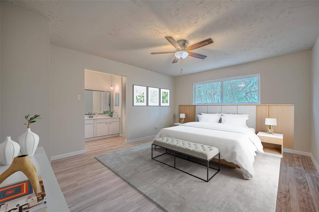 This is a spacious, well-lit primary bedroom featuring a modern ceiling fan. The room has a neutral color, wood-like flooring, and offers a view into an en suite bathroom and the outdoors through a large window.