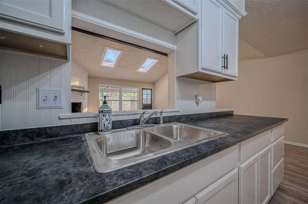 This kitchen features white cabinetry, dark countertops, a stainless steel double sink, and natural light from skylights. A pass-through window overlooks the living area with a fireplace.